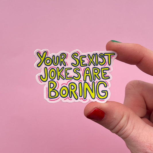 Hand holding a sticker with 'Your Sexist Jokes Are Boring' text against a pink background