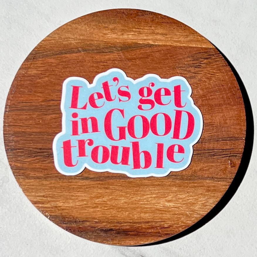 Round wooden coaster with a colorful sticker reading 'Let's get in Good trouble' on a light gray background.