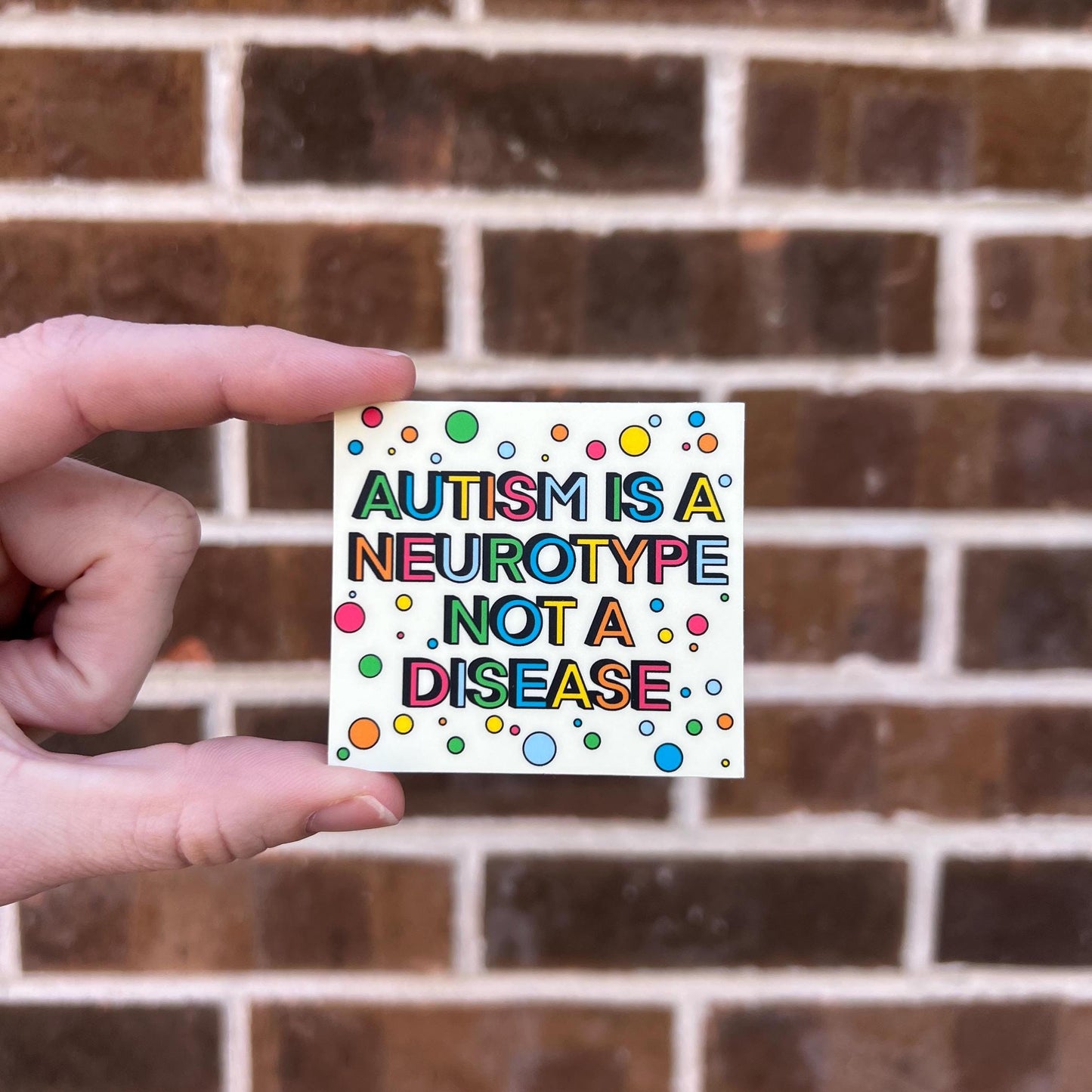 hand holding a sticker that says "autism is a neurotype not a disease" on a brick background