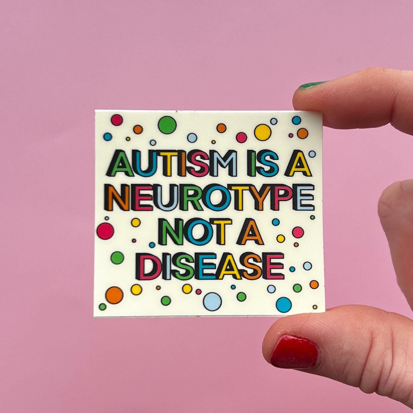 hand holding a sticker that says "autism is a neurotype not a disease" on a pink background