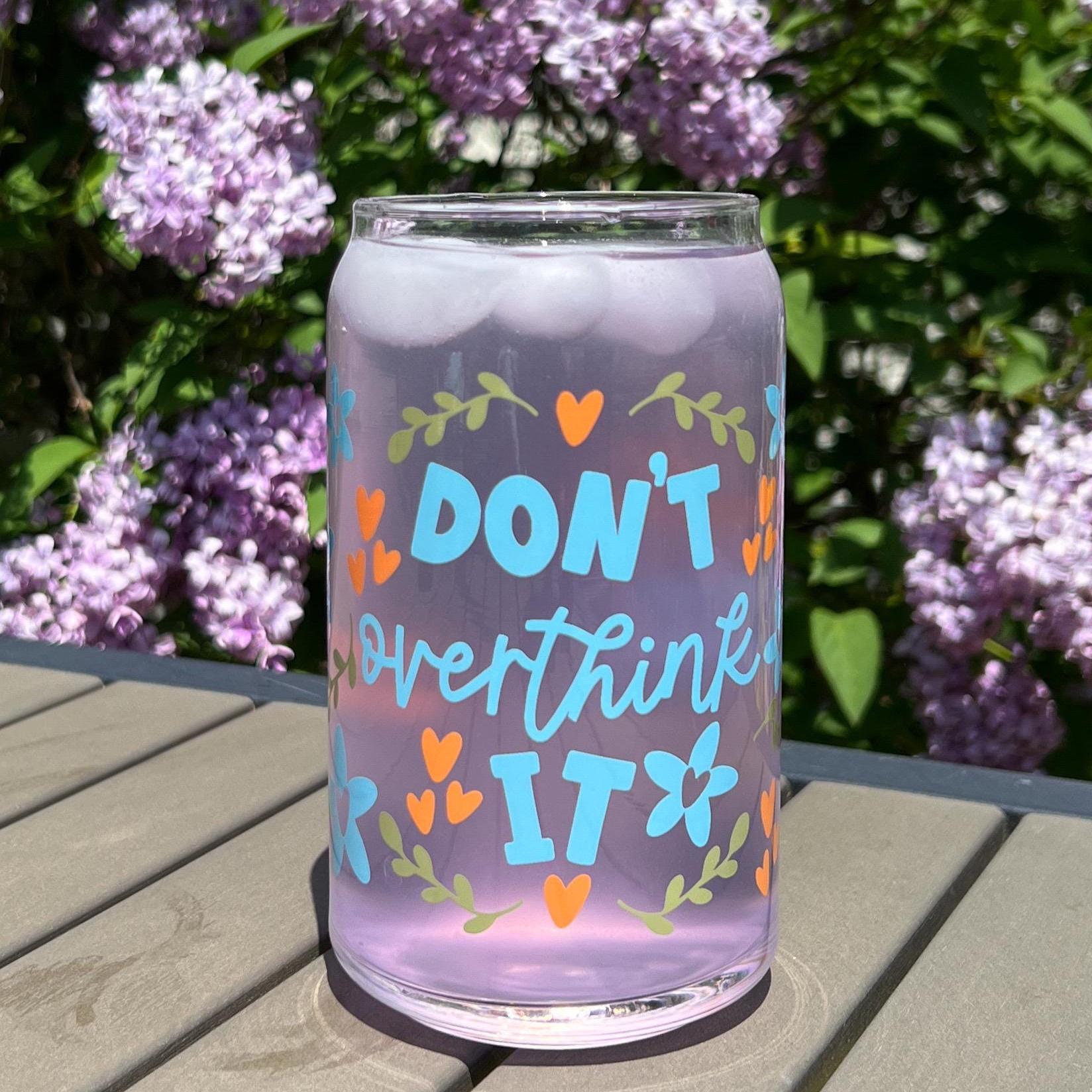 Glass tumbler with 'Don't overthink it' text on a wooden surface with purple flowers in the background