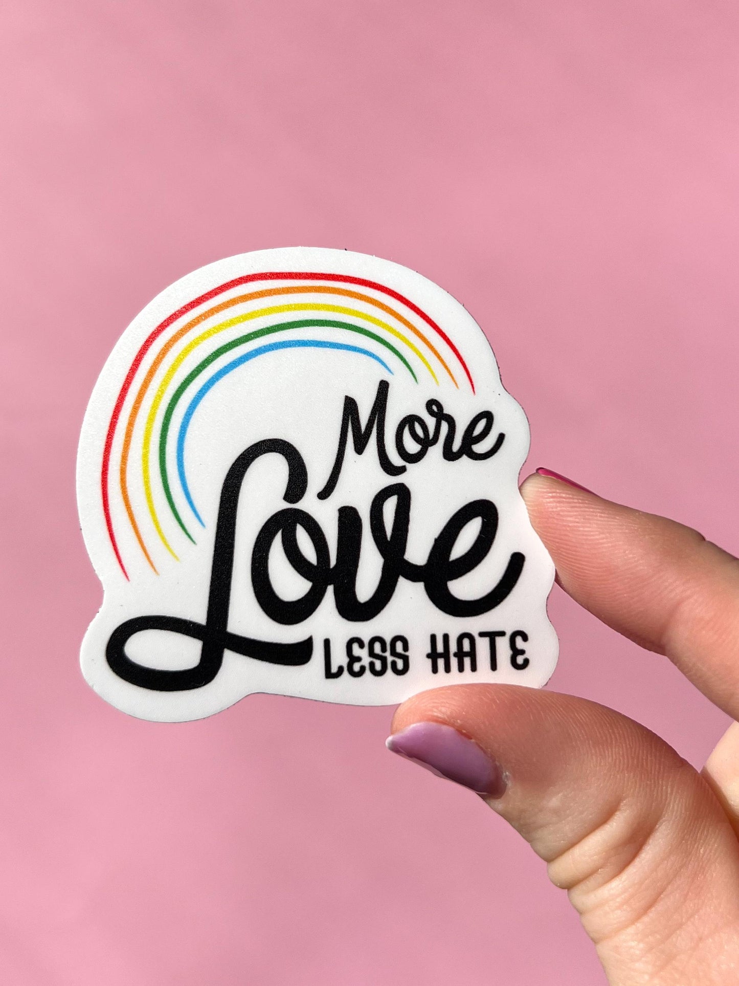 Sticker with rainbow and text 'More Love Less Hate' held by a hand against a pink background