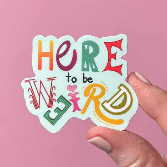 Colorful sticker with 'Here to be WEIRD' text held by a hand against a pink background